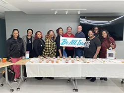 Image of a group of people standing behind a table, holding a sign that says be all in