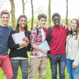 Five teenagers standing in a line, in an outdoor setting, with arms wrapped around one another, smiling at the camera. 