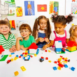 5 children, with two boys on the left and three girls on the right, are playing with Legos at a white table with artwork hanging up in the background. 