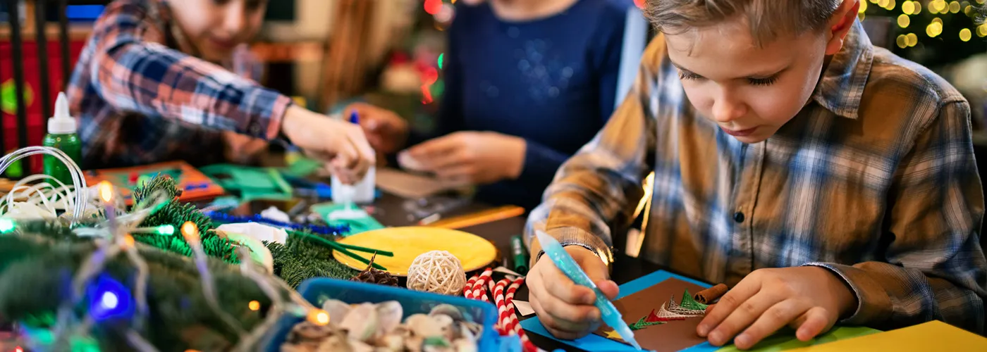 Kids crafting holiday greeting cards with festive lights in the background