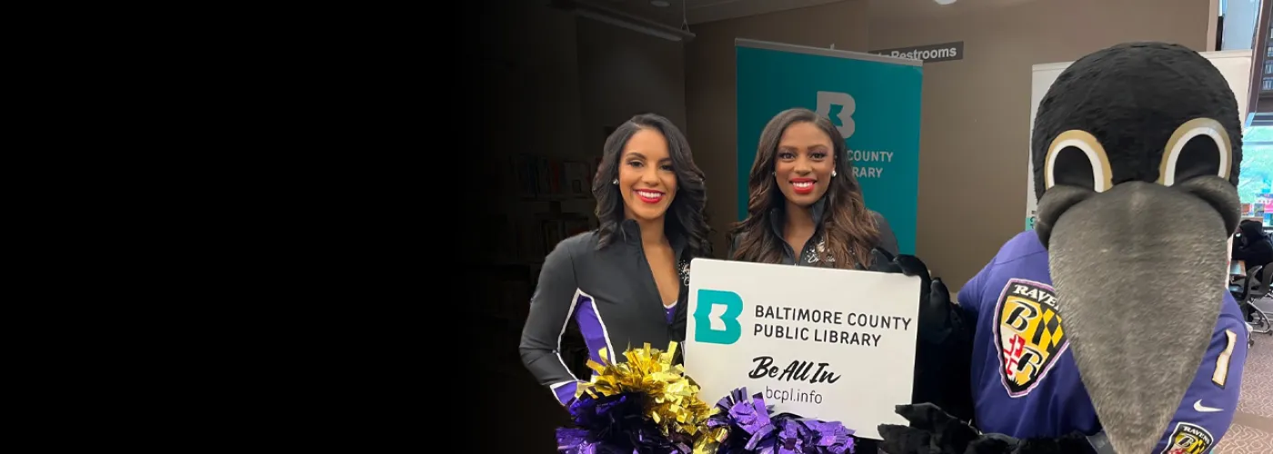 The Baltimore Ravens Mascot Poe and two cheerleaders posing with an oversized library card